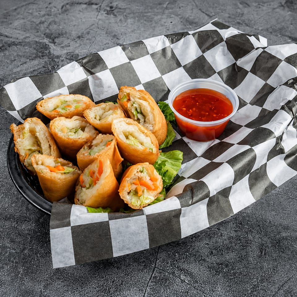 Crispy fried spring rolls, cut in half, with sweet chili dipping sauce in a checkered paper-lined basket.