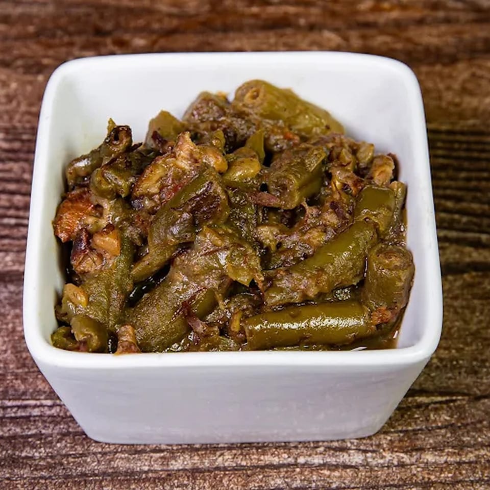 A white square bowl filled with cooked green beans and meat on a wooden surface.