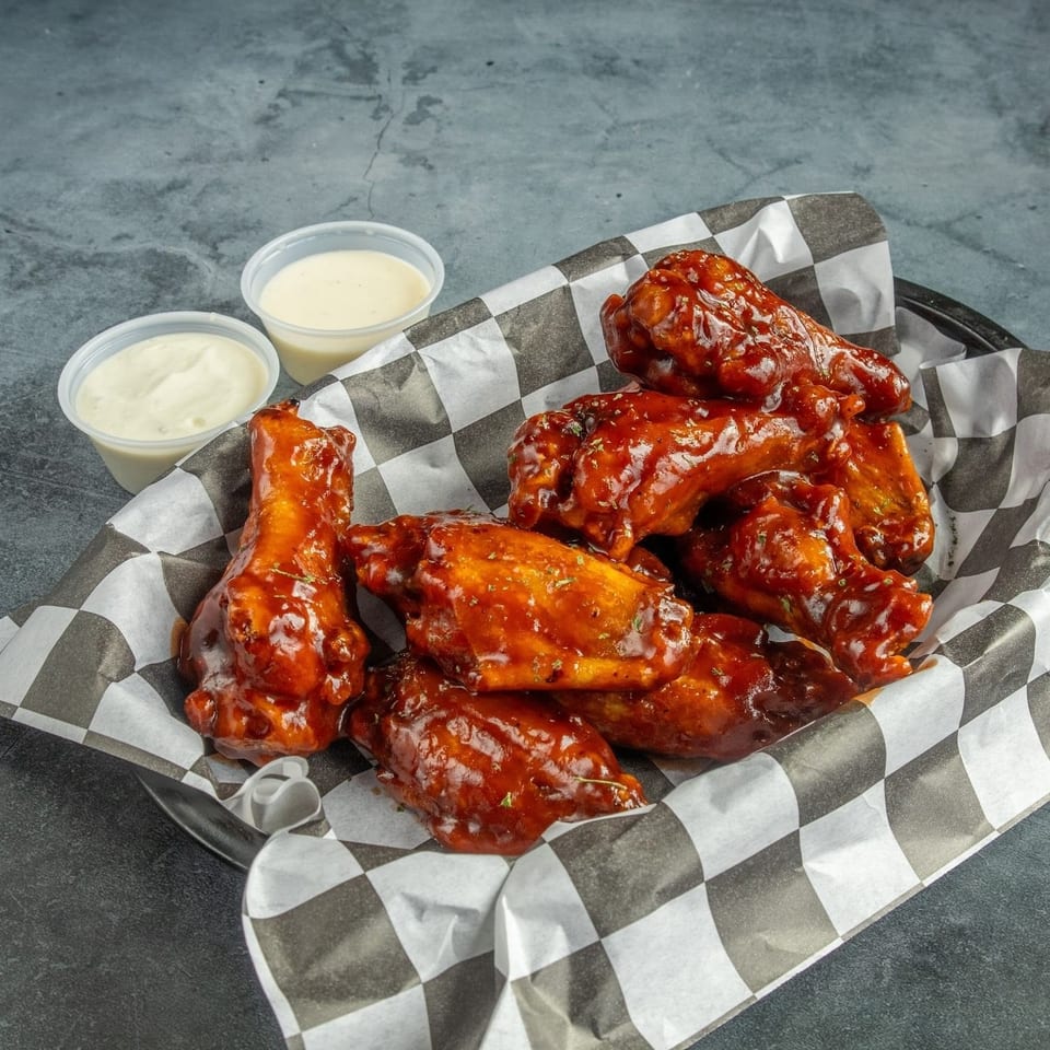 A basket of saucy chicken wings with two dipping sauces on a dark gray surface.