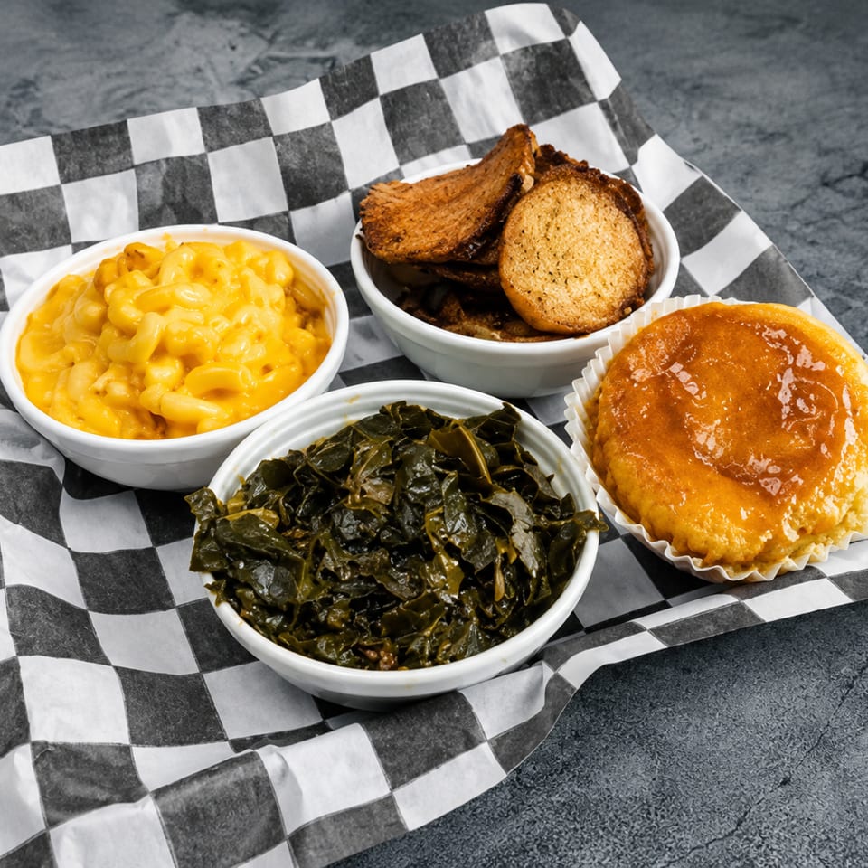 Macaroni and cheese, collard greens, cornbread, and hush puppies on checkered paper.