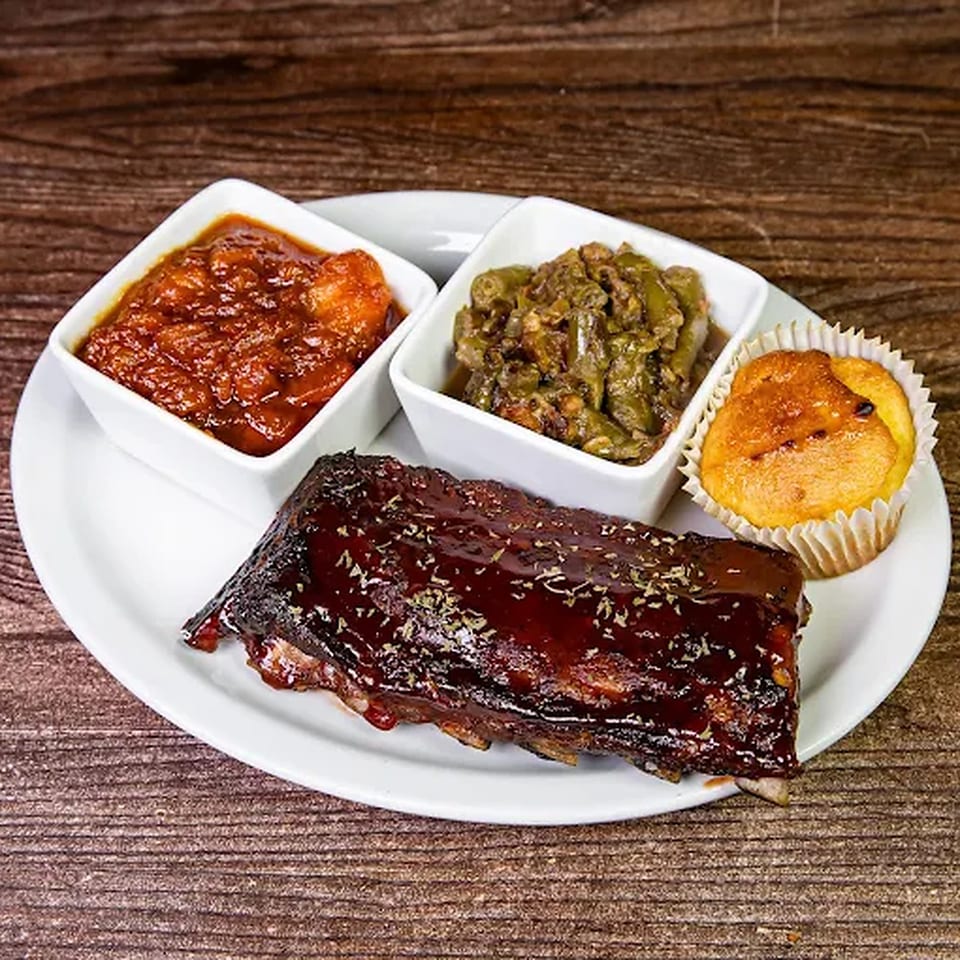 A plate of BBQ ribs with two side dishes and a cornbread muffin on a wooden table.